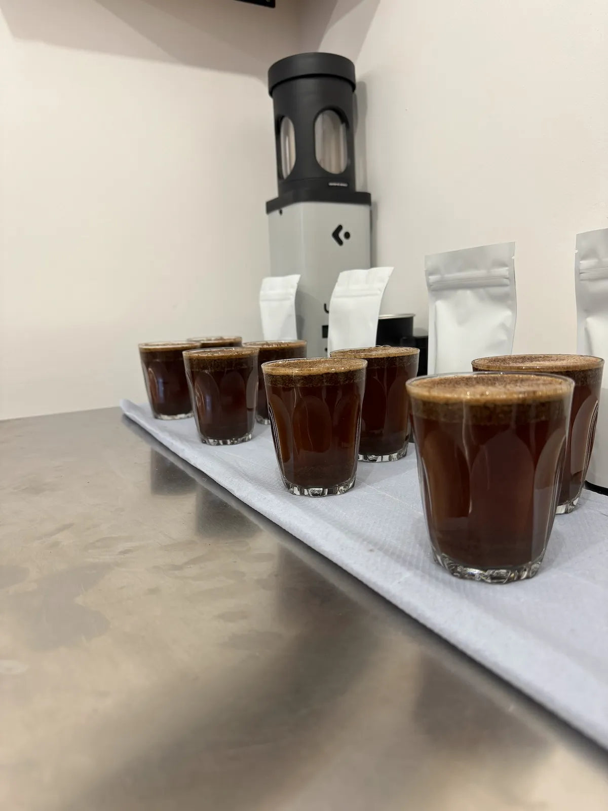Row of coffee samples in glasses on a metal surface with coffee bags in the background.